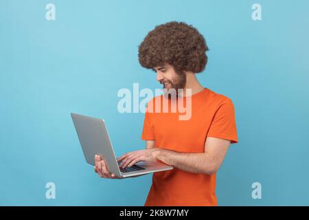 Portrait of man with Afro hairstyle in orange T-Shirt hält Laptop in der Hand und tippt, Blogger macht Beiträge in sozialen Netzwerken, chattet mit Followern. Innenaufnahme des Studios isoliert auf blauem Hintergrund. Stockfoto