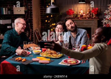 Frau, die am festlichen weihnachtstisch ein Porträtfoto von verschiedenen Menschen gemacht hat. Großes Familientreffen, Frau und Ehemann feierten Weihnachten mit den Eltern und tranken Sekt Stockfoto