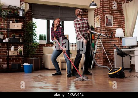 Frau wischt den Boden und tanzt, während der Mann mit Leidenschaft mit dem Staubsauger singt. afroamerikanisches Paar putzt am Wochenende das Haus und sorgt dafür, dass das Haus ordentlich und sauber bleibt. Stockfoto