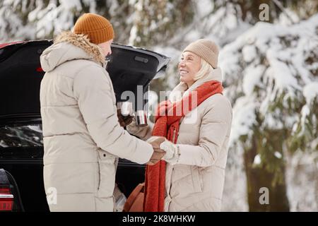 Glückliches Seniorenpaar genießt eine Tasse heißen Kokos im Freien im Winterwald und hält sich die Hände Stockfoto