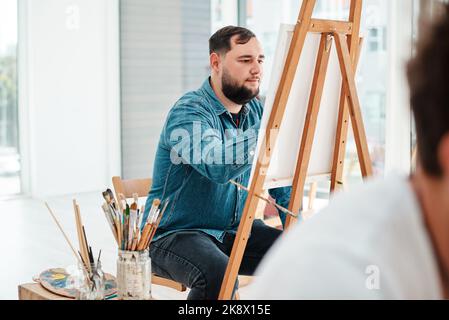 Ich kann durch Kunst kreativ sein. Ein hübscher junger Künstler, der während eines Kunstuntermalens im Atelier sitzt und malt. Stockfoto