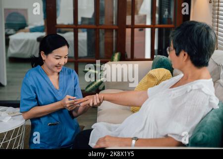 Positive asiatische weibliche Pflegekraft in blauen Peelings lächelnd und massierenden Fingern einer älteren Frau, die während der Reha-Sitzung zu Hause auf dem Sofa sitzt Stockfoto