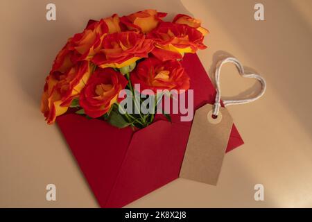 Schöne rote Rosen Blumen in postalischem roten Umschlag auf rotem Hintergrund, beige Papiernote und Seil in Herzform Kopieplatz für Text, Frühling, Grußkarte für Urlaub. Blumenlieferung. Zarte rote gelbe Rosen. Monochrome Minimal trendige Komposition. Romantische Blumen Stockfoto