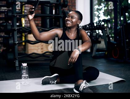Selfie im Fitnessstudio. Ganzkörperaufnahme einer attraktiven jungen Sportlerin, die ein Selfie macht, während sie im Fitnessstudio auf ihrem Handtuch sitzt. Stockfoto