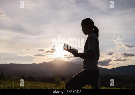 Silhouette einer Frau, die zu gott betet, mit der bibel auf dem Berg im Hintergrund des Sonnenuntergangs. Frau, die seine Hände in Anbetung hob. Christliches Religionskonzept. Stockfoto