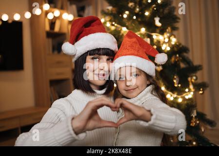 Nahaufnahme der schönen Familie von zwei, schöne junge Mutter und ihre niedliche kleine Tochter in warmen gestrickten weißen Pullover machen herzförmige Zeichen mit ihren Händen vor verzierten Weihnachtsbaum. Stockfoto