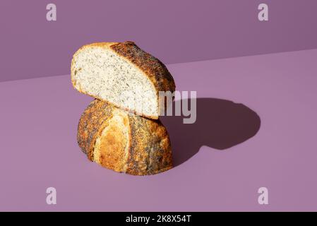 Frisch gebackenes Brot mit Mohnsamen, minimalistisch auf einem violetten Tisch. Hausgemachtes Brot in zwei Hälften geschnitten in hellem Licht vor einem bunten Hintergrund Stockfoto