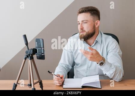 Lächelnder bärtiger Geschäftsmann, der seine Video-Blog-Episode mit dem Smartphone filmt, am Schreibtisch sitzt, schreibt in ein Notebook und gestikuliert mit seiner Hand. In einem Hemd gekleidet Stockfoto
