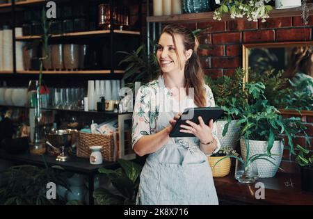 Ich bin bereit für einen anstrengenden Tag. Eine attraktive junge Geschäftsfrau, die allein in ihrem Blumenladen steht und ein Tablet benutzt. Stockfoto