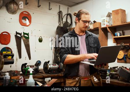 Ich muss mich das Design noch einmal ansehen, bevor ich weitermache. Ein fokussierter junger Zimmermann, der an seinem Laptop arbeitet, während er in seiner Werkstatt steht Stockfoto