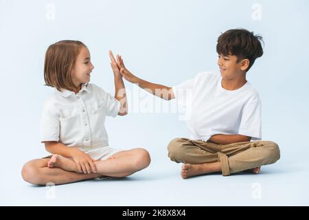 High Fiving Junge und Mädchen sitzen auf dem Boden mit gekreuzten Beinen, Blick auf ihre Hände. Gegen bläulich weißen Hintergrund. Beide barfuß. Stockfoto