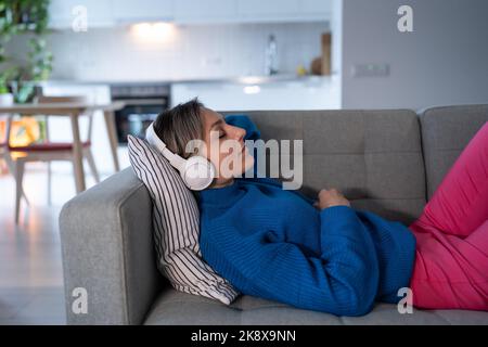 Junge Frau mit weißen Kopfhörern hört gerne ruhige Musik und ruht sich auf einem bequemen Sofa aus Stockfoto