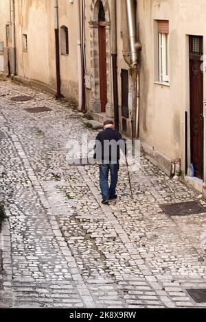 Älterer Mann mit coppola Hut und Gehstock auf der Steinstraße des mittelalterlichen Dorfes Prizzi in Westsizilien, Italien Stockfoto