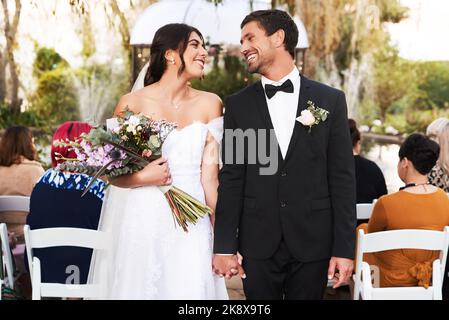 Du bist alles, was ich in dieser Welt brauche. Ein glückliches junges Paar posiert zusammen im Freien an ihrem Hochzeitstag mit Gästen im Hintergrund. Stockfoto