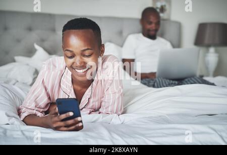 Jeder hat ein Lieblingsgerät. Eine glückliche junge Frau benutzt ein Smartphone, während ihr Mann zu Hause einen Laptop im Bett benutzt. Stockfoto