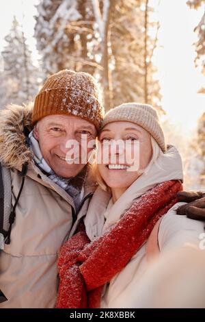 Vertikaler Blickwinkel eines glücklichen älteren Paares, das Selfie-Fotos macht, während er eine Wanderung im Winterwald genießt Stockfoto