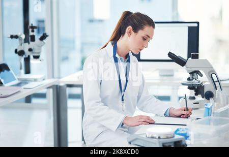 Ich muss ein paar Notizen zu diesem Thema machen. Eine fokussierte junge Wissenschaftlerin, die tagsüber Tests macht und Notizen in einem Labor macht. Stockfoto