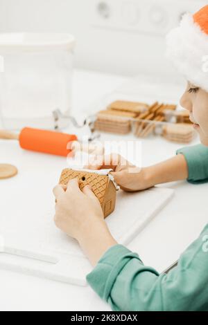 Ein süßer Junge an Silvester und Weihnachten macht in einem Weihnachtshut ein Ingwerhaus in der Küche. Stockfoto