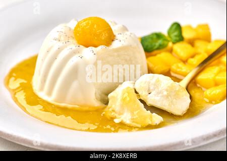 Makrolöffel mit Stück hausgemachter Panna Cotta mit natürlicher Vanilleschote, Mangosirup und Minze, selektiver Fokus Stockfoto