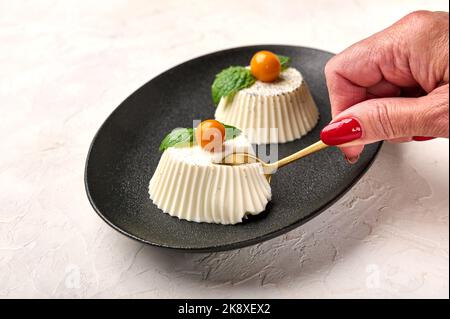 Frauenhand mit Löffel bricht ein Stück hausgemachte Panna Cotta mit natürlicher Vanilleschote, Physalis oder Winterkirsche und Minze auf schwarzem Teller ab, selektiver Fokus Stockfoto