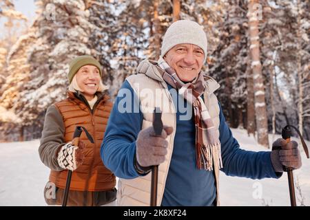 Porträt eines glücklichen älteren Paares, das mit Stöcken im Winterwald spazierengeht und die Kamera anlächelt Stockfoto