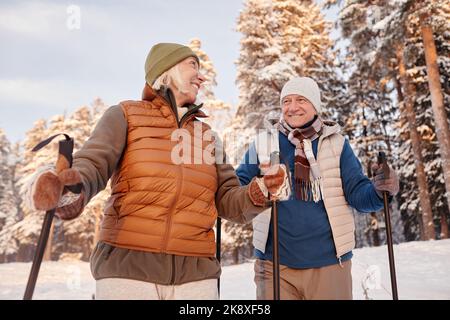 Porträt eines glücklichen älteren Paares, das mit Stöcken im Winterwald spazierengeht und lächelt Stockfoto