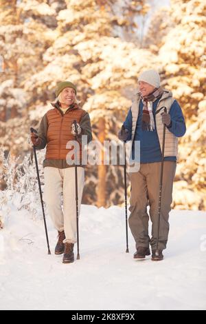 Porträt eines aktiven älteren Paares, das mit Stöcken in einem wunderschönen Winterwald spazierengeht und lächelt Stockfoto