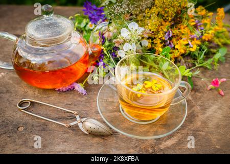 Sommerlicher gesunder Kräutertee in einer Glas-Teekane und Kräuterbündel auf Holzbrett. . Phytotherapie. Kräutermedizin Stockfoto