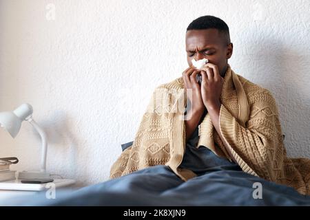 Ein hübscher junger Mann sitzt in seinem Bett und bläst seine Nase, während er an einer Erkältung leidet. Stockfoto