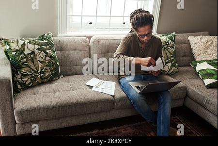 Bezahlung der Rechnungen vor den Feiertagen. Ein fokussierter junger Mann, der an einem Laptop arbeitet und Papierkram macht, während er tagsüber auf einem Sofa zu Hause sitzt. Stockfoto