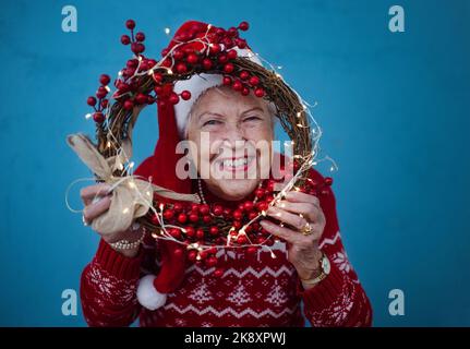 Porträt einer glücklichen älteren Frau mit Weihnachtsmannmützen im Studio, die durch den Weihnachtskranz schaut. Stockfoto