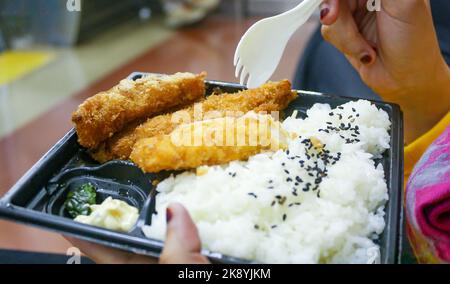 Nahaufnahme der Hände mit Kunststoffplatte und Löffel. Frittiertes Hähnchenschnitzel und Reis mit schwarzem Sesamsamen auf einem Teller mit Bokeh-Hintergrund. Stockfoto