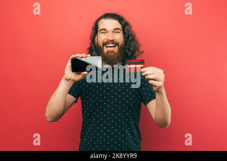 Foto des glücklichen bärtigen Mannes yong mit langen Haaren und Smartphone-Bildschirm und roter Debitkarte. Stockfoto