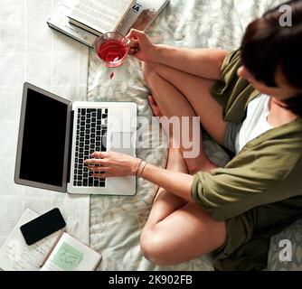 Online nur die neuesten Trends nachholen. Aufnahme einer jungen Frau, die Tee trinkt und einen Laptop benutzt, während sie sich zu Hause in ihrem Schlafzimmer entspannt. Stockfoto