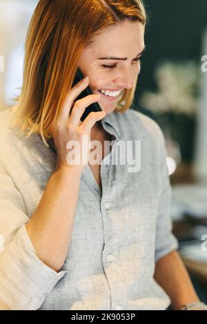 Glückliche junge Geschäftsfrau, die fröhlich lächelt, während sie während eines Telefonanrufs spricht. Erfolgreiche Geschäftsfrau, die mit ihren Geschäftspartnern in einem kommuniziert Stockfoto