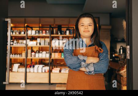 Selbstbewusste Frau mit Down-Syndrom, die am Eingang eines Delikatessenessenläden steht. Ermächtigte Frau mit einer intellektuellen Behinderung, die als Ladenbesitzerin in einem lo arbeitet Stockfoto