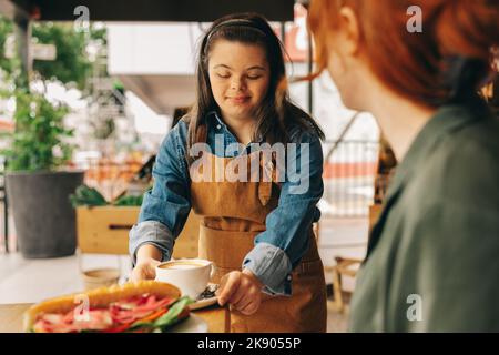 Waitron mit Down-Syndrom serviert einem Kunden ein Sandwich und Kaffee in einem trendigen Café. Professionelle Frau mit einer intellektuellen Behinderung, die in einem Stockfoto