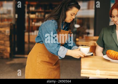 Professionelle Kellnerin mit Down-Syndrom serviert einem Kunden ein Sandwich und Kaffee in einem trendigen Café. Frau mit einer intellektuellen Behinderung, die in einem Stockfoto