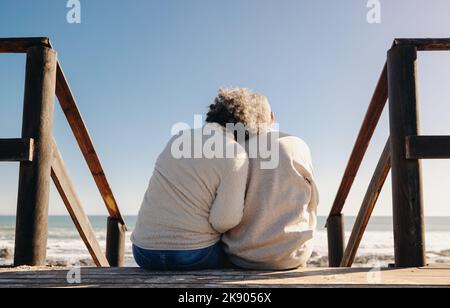 Rückansicht eines älteren Paares, das einen erfrischenden Blick auf das Meer hat, während es auf einer hölzernen Fußbrücke sitzt. Rentnerpaar, das etwas Qualität ausgegeben hat Stockfoto