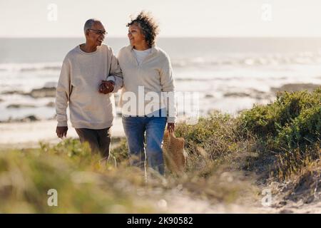 Reifes Paar lächelt fröhlich, während es nach einem romantischen Picknick vom Strand weg geht. Fröhliches, älteres Paar, das nach dem Urlaub am Meer einen Urlaub genießt Stockfoto