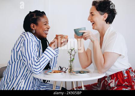 Zwei glückliche Geschäftsfrauen, die lachen und gemeinsam in einem Bürocafé Kaffee trinken. Zwei fröhliche Geschäftsfrauen genießen ein freundliches Kaffeetreffen in einem Mod Stockfoto
