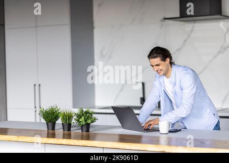 Serious fokussierten bestimmten jungen bärtigen Geschäftsmann oder Freiberufler in Gläsern und Freizeitkleidung sitzen am Küchentisch, tippen auf Laptop, arbeiten o Stockfoto