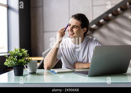 Ein nachdenklicher Geschäftsmann, der eine Brille trägt, die das Kinn berührt, Ideen oder Strategien nachdenkt, am Holzarbeitstisch mit Laptop sitzt und freiberuflich an Onli arbeitet Stockfoto