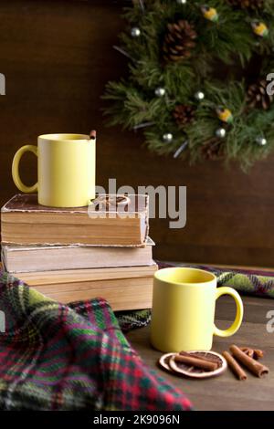 Bücher und zwei gelbe Tassen in der Nähe des Kranzes mit Zimt in der Nähe von weihnachtsdekorationen auf einem Holztisch Stockfoto