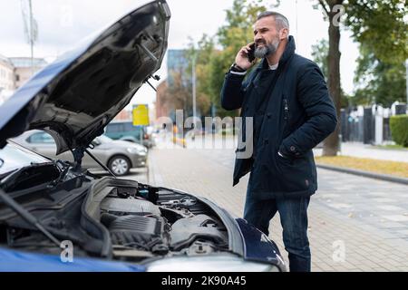 Ein erwachsener bärtiger grauhaariger Mann spricht auf einem Mobiltelefon in der Nähe der offenen Haube eines kaputten Autos Stockfoto