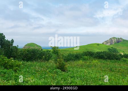 Natürliche Landschaft der Insel Kunashir mit grasbewachsenen Hügeln, vulkanischen Felsen, Vulkan in den Wolken und Ozean in der Ferne Stockfoto