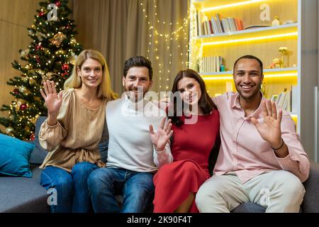 Portrait der Neujahrsparty mit diversen Freunden an Weihnachten, Gäste, die auf die Kamera schauen, winkende Grüße und lächelnd, sitzen auf dem Sofa in der Nähe des Weihnachtsbaums im Wohnzimmer. Stockfoto