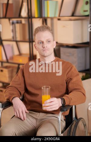 Porträt eines nachdenklichen jungen Mannes, der im Rollstuhl sitzt, ein Glas Saft trinkt und über das Leben nachdenkt Stockfoto