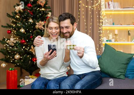 Verheiratetes Paar in Liebe Weihnachtseinkäufe entfernt im Online-Shop, glücklicher Ehemann und Ehefrau sitzen auf dem Sofa in der Nähe des Weihnachtsbaums, halten Bankkreditkarte und Telefon in den Händen und feiern das neue Jahr. Stockfoto