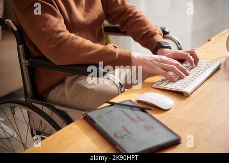 Zugeschnittenes Bild eines jungen Mannes im Rollstuhl, der Modeskizzen zeichnete und E-Mails von Kunden beantwortete Stockfoto